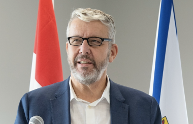 Man in a blue blazer with white shirt and black glasses stands at a microphone. The man stands in front of a Canadian flag and Nova Scotia flag.