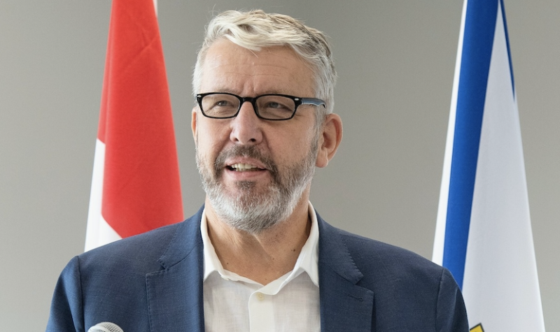 Man in a blue blazer with white shirt and black glasses stands at a microphone. The man stands in front of a Canadian flag and Nova Scotia flag.