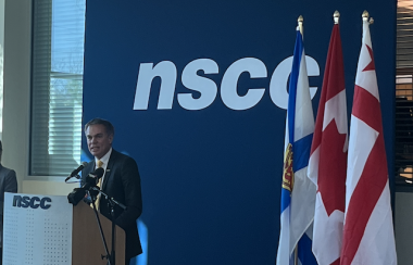 Man stands behind a podium in front of a dark blue backdrop with NSCC written on it. Also in the picture are Nova Scotia, Canadian and Mi'kmaw flags.