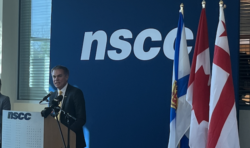 Man stands behind a podium in front of a dark blue backdrop with NSCC written on it. Also in the picture are Nova Scotia, Canadian and Mi'kmaw flags.