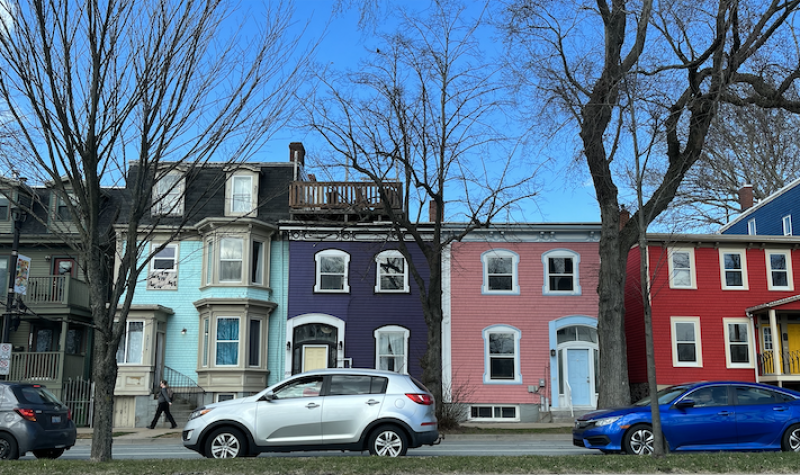 Row of colourful houses with cars parked on the street in front. Houses are blue, purple, pink and red.