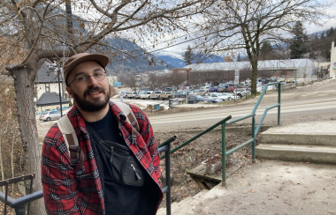 James Barbeiro is facing the camera, dressed in a red shirt and brown hat with nelson bc mountains in the background