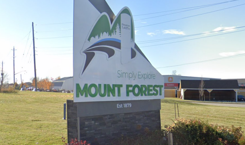 A sunny day in Mount Forest. A sign for Mount Forest sits on a lawn reading 'Simply Explore.'