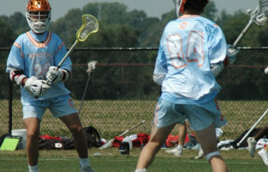 Two lacrosse players in orange helmets and powder blue jerseys, Blake Willard and Riley Woods, compete in the Platinum Cup.