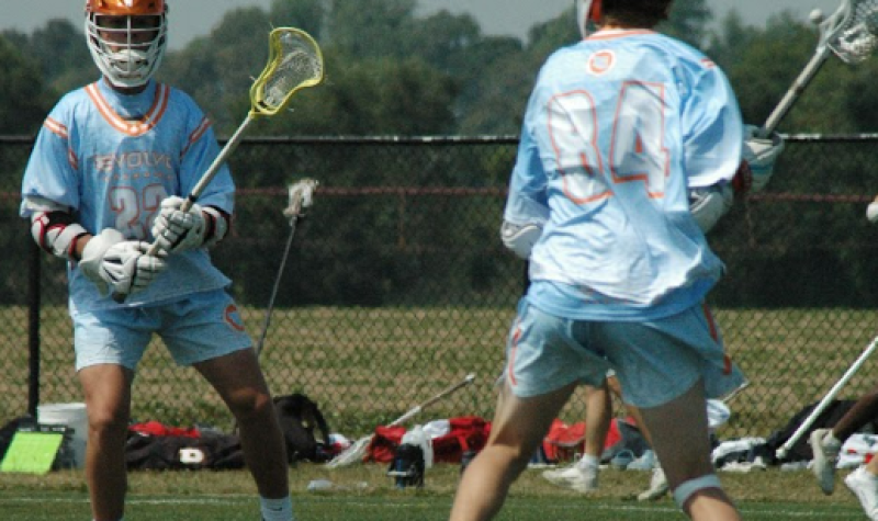 Two lacrosse players in orange helmets and powder blue jerseys, Blake Willard and Riley Woods, compete in the Platinum Cup.