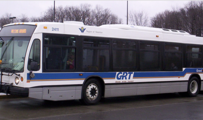 a white, blue and grey bus is parked at a bus stop next to a bus shelter on a grey, rainy day.