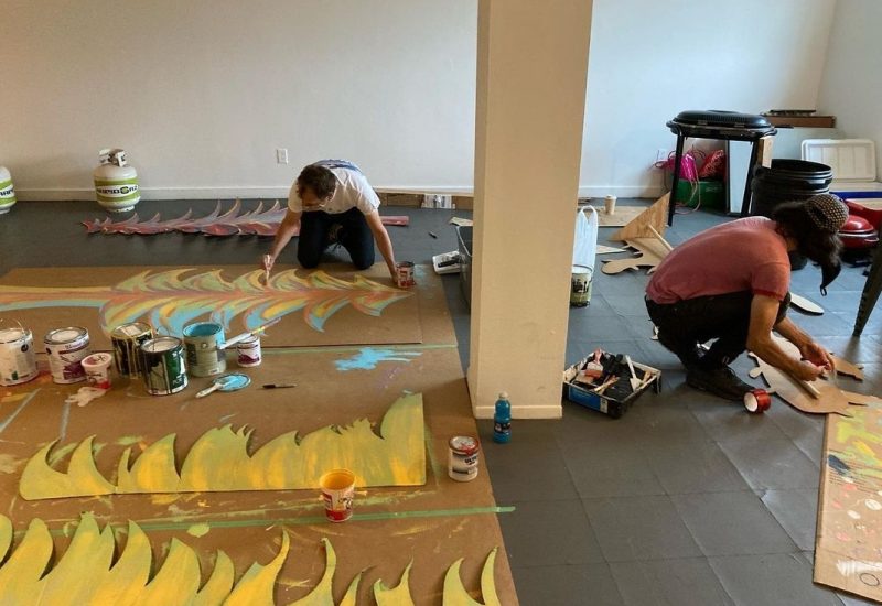 Two people kneel on the floor and paint yellow patterns on brown paper.