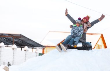 Femme habillée d’un habit de neige avec un enfant sur ses genoux qui glissent dans un tube pour la neige. Ils sont en hauteur et l’on aperçoit un immeuble à l’arrière.