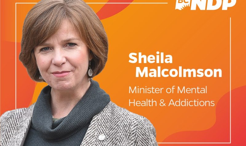Sheila Malcolmson in front of orange banner with her name and position as minister