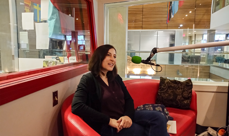 A woman with shoulder length dark hair, Shelley Liebembuk, sits on a red couch with a microphone to do record an interview with station hosts and UFV CHASI members.