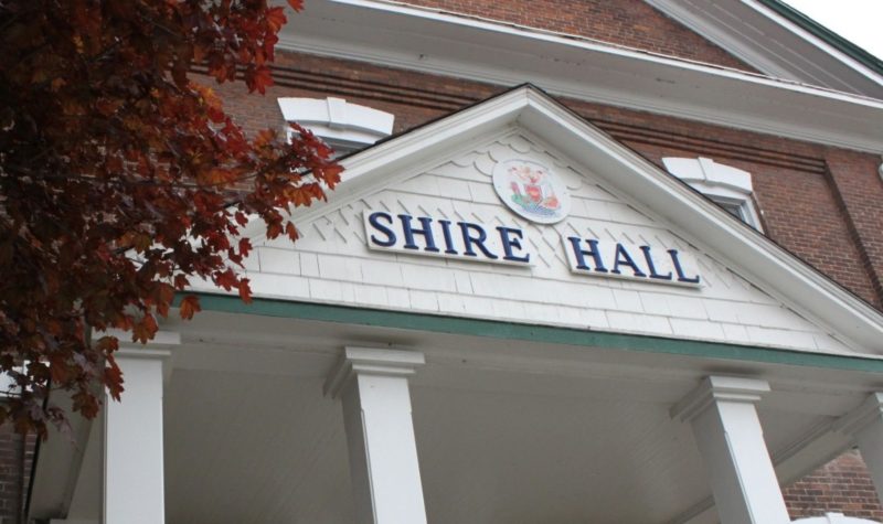 A two story brick building with a white peaked entryway with four columns. The front of the building says 'Shire Hall'.
