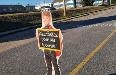 Silhouette flexible d'un enfant garçon tenant une affiche écrit: Ralentissez pour ma sécurité sur une rue avec les écoles de quartier derrière.