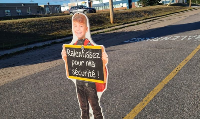 Silhouette flexible d'un enfant garçon tenant une affiche écrit: Ralentissez pour ma sécurité sur une rue avec les écoles de quartier derrière.