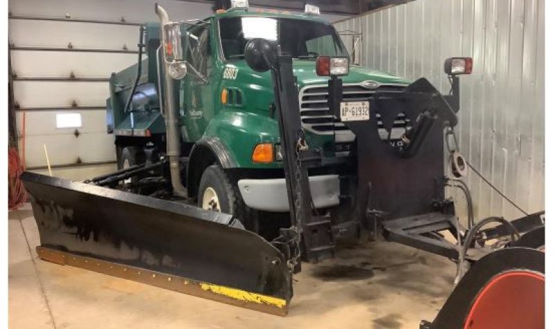 A large green dump truck inside a white and metallic garage structure. The truck has a large side shovel attached to its frontal section.