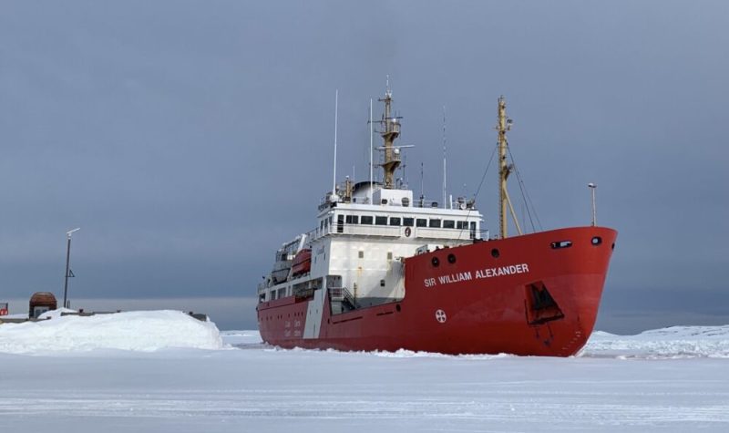 Un navire à la cale rouge aux abords d'un quai. Il semble reposer sur un sol enneigé et glacé.