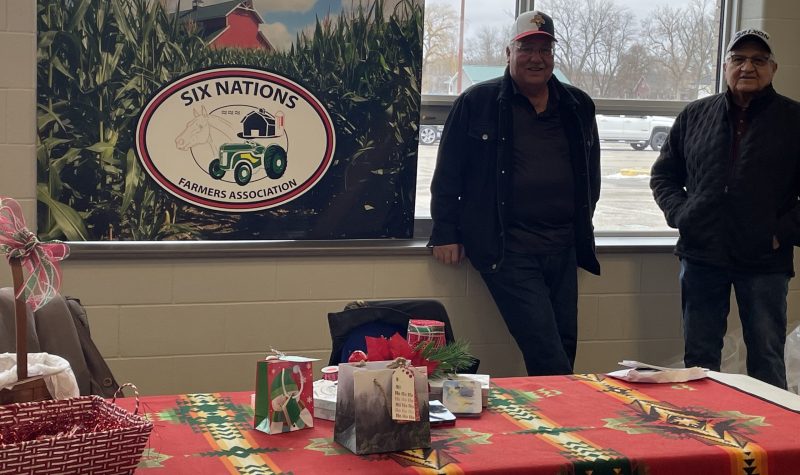 two men stand against window with Farmers sign beside them and table infront draped with blanket and items