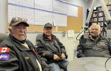Three men sit around a round table in a radio studio, all wearing black jackets.