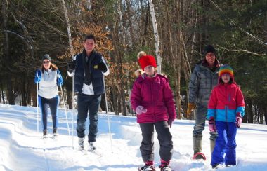 A group of adults and children cross-country skiing and snowshoeing on a wooded trail.