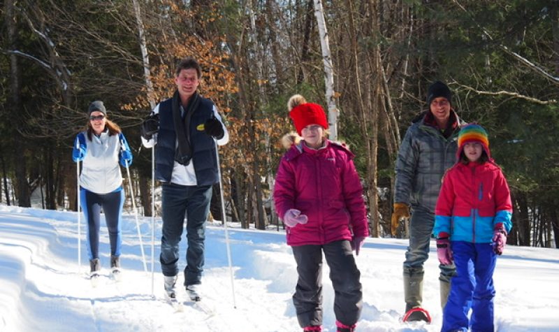 A group of adults and children cross-country skiing and snowshoeing on a wooded trail.