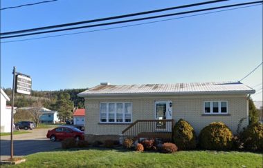 Petite maison beige avec la porte et les fenêtres blanches. Petite galerie en bois devant et verdures devant la maison. Il y a une enseigne à gauche inscrite : Société Financière du Saguenay ltée.