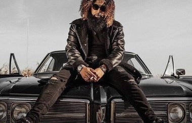 L'artiste 4Say cheveux et barbe bouclés, lunettes noires, veston en cuir, assis sur le capot d'une voiture noire