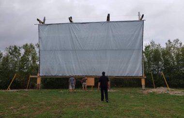 A large white movie drive-in screen is seen in a field with a man standing in front of it looking up at it on an overcast day.