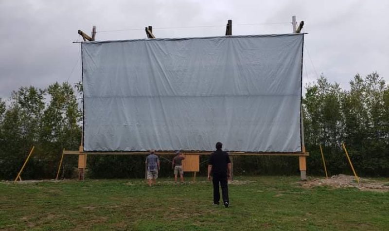 A large white movie drive-in screen is seen in a field with a man standing in front of it looking up at it on an overcast day.