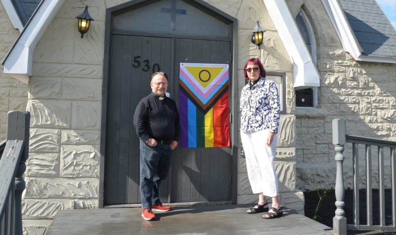 Two Anglican Reverends stand in front of their church with a Pride flag attached to the door.