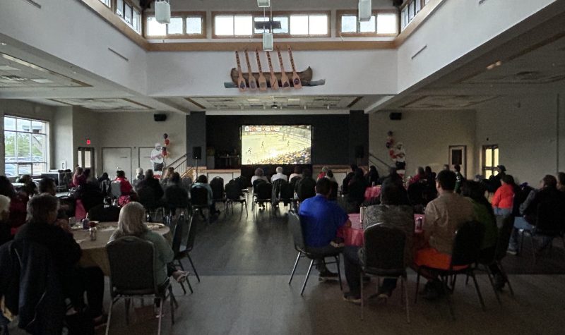 A darkened room full of people sit watching a large screen of the Stanley cup Final at Six Nations