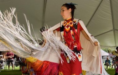 On aperçoit une femme habillée avec des vêtement traditionnels autochtones performant une danse sous un chapiteau installé sur le site historique de Ste-Marie-au-pays des-Hurons. Derrière l'artiste, des visiteurs observent la performance.