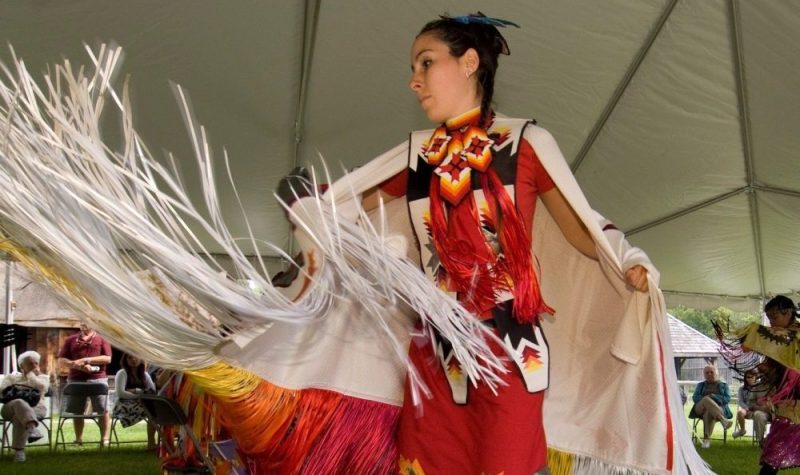 On aperçoit une femme habillée avec des vêtement traditionnels autochtones performant une danse sous un chapiteau installé sur le site historique de Ste-Marie-au-pays des-Hurons. Derrière l'artiste, des visiteurs observent la performance.