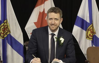 Iain Rankin writes in a book at a podium at a press conference with Nova Scotia flags behind him