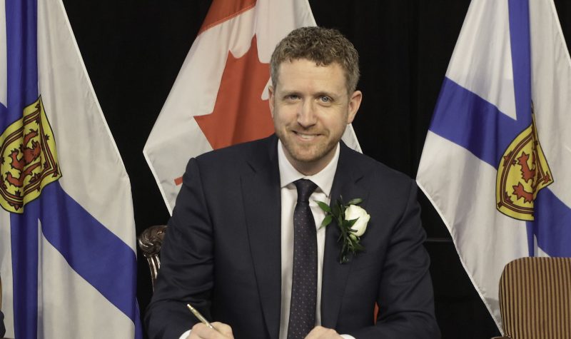 Iain Rankin writes in a book at a podium at a press conference with Nova Scotia flags behind him