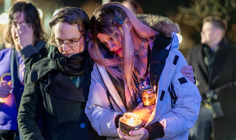 Two people in winter jackets comfort one another with one person holding a candle.