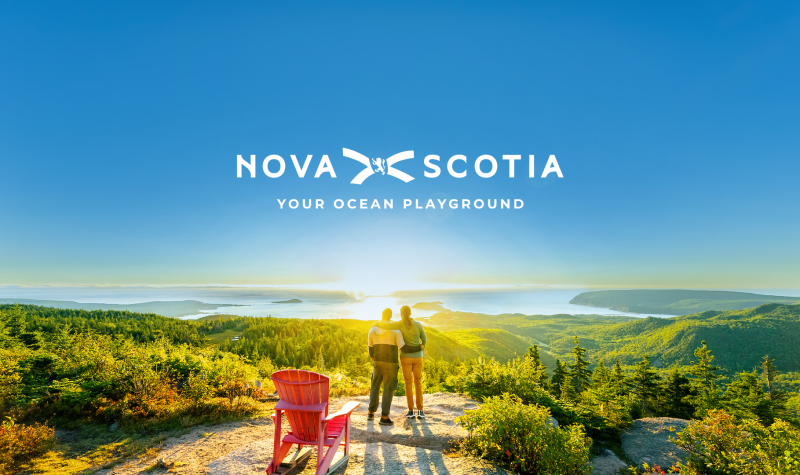 Photo of the latest Nova Scotia tourism campaign that shows a couple standing over a long shoreline. The graphic text says Nova Scotia Your Ocean Playground.
