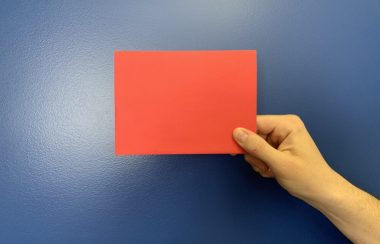 Une main tient un carton rouge devant un mur bleu.
