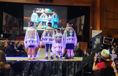 Four boys on a stage with their backs to the camera showing traditional indigenous designs on white capes