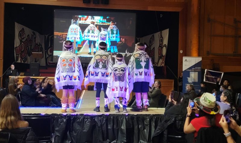 Four boys on a stage with their backs to the camera showing traditional indigenous designs on white capes