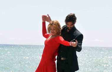A woman in red dress in dance postions with man in black suit by the shore