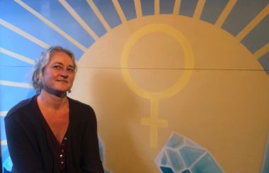 Cortes Island Women's Resource Centre Founder and Director Tanya Henck stands in front of a large painting of the centre's logo with yellow and blue graphics.