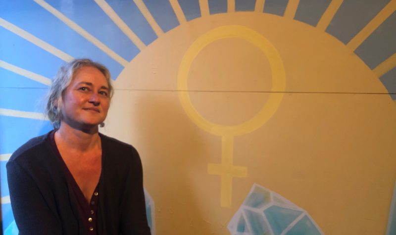 Cortes Island Women's Resource Centre Founder and Director Tanya Henck stands in front of a large painting of the centre's logo with yellow and blue graphics.