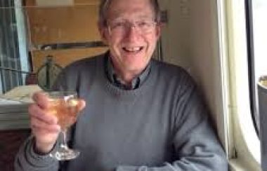 A man is seen on a train holding up a glass of white wine.