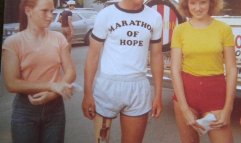 Terry Fox taking a photo with two young women in a 1980s photo. He is wearing a white t-shirt and white shorts and they are standing in a parking lot.