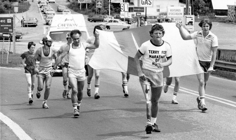 A black and white dated photo of Terry Fox running on a road with several other behind him and carrying a flag