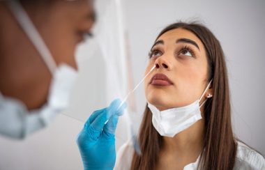 Femme aux cheveux bruns, portant un masque de procédure sous le menton, se fait faire un test PCR par une professionnelle.