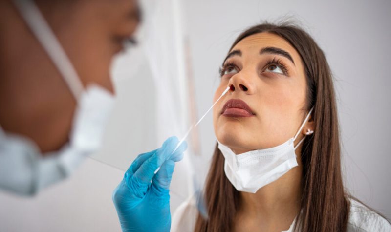 Femme aux cheveux bruns, portant un masque de procédure sous le menton, se fait faire un test PCR par une professionnelle.