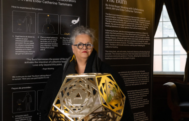 A person with glasses stands behind a golden object that is on a stand. There are black, long sheets with white text behind the person.