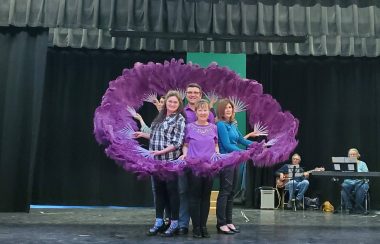 Four people on a stage surrounded by purple feathers rehearse a play.
