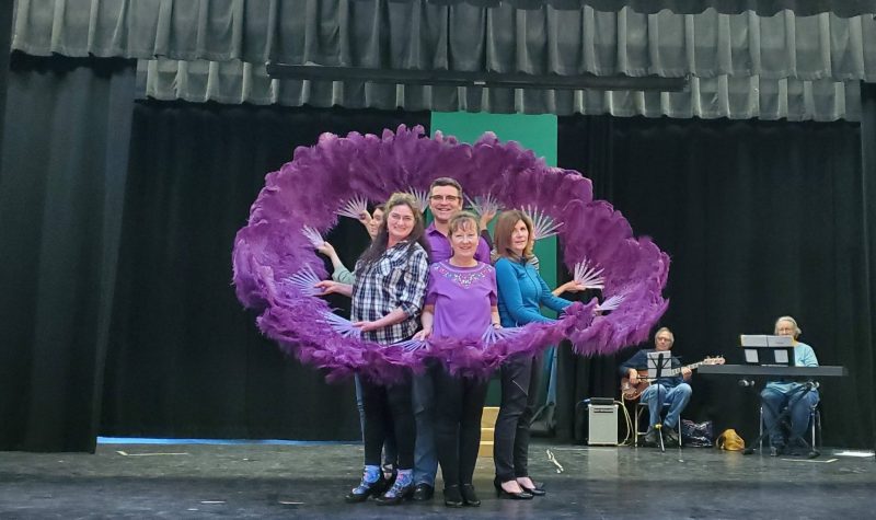 Four people on a stage surrounded by purple feathers rehearse a play.