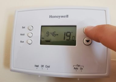 A white Honeywell thermostat on the wall with a finger pressing a button.